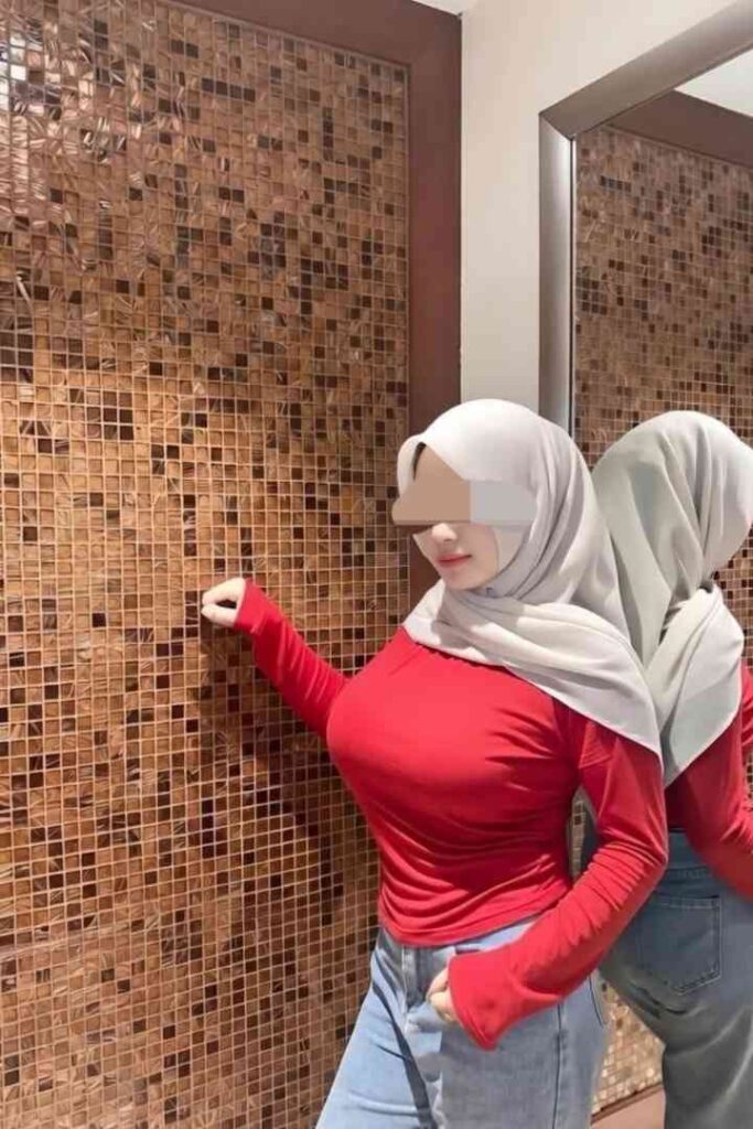 Woman in red top and hijab posing by tiled wall and mirror, reflecting her image.