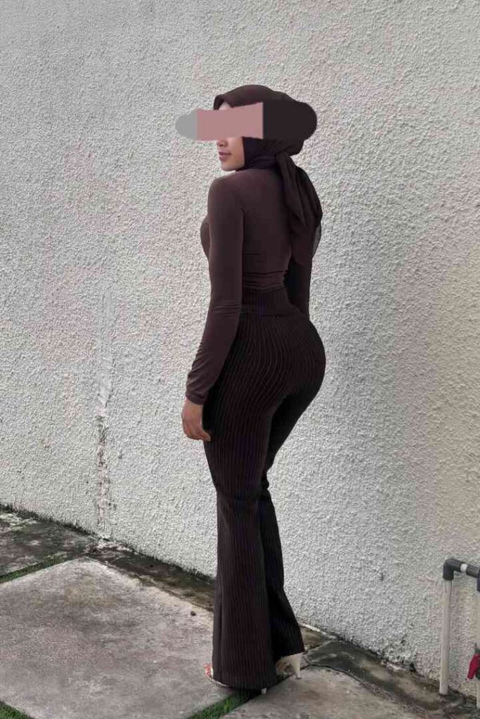 Woman in brown hijab and outfit standing against a textured wall.